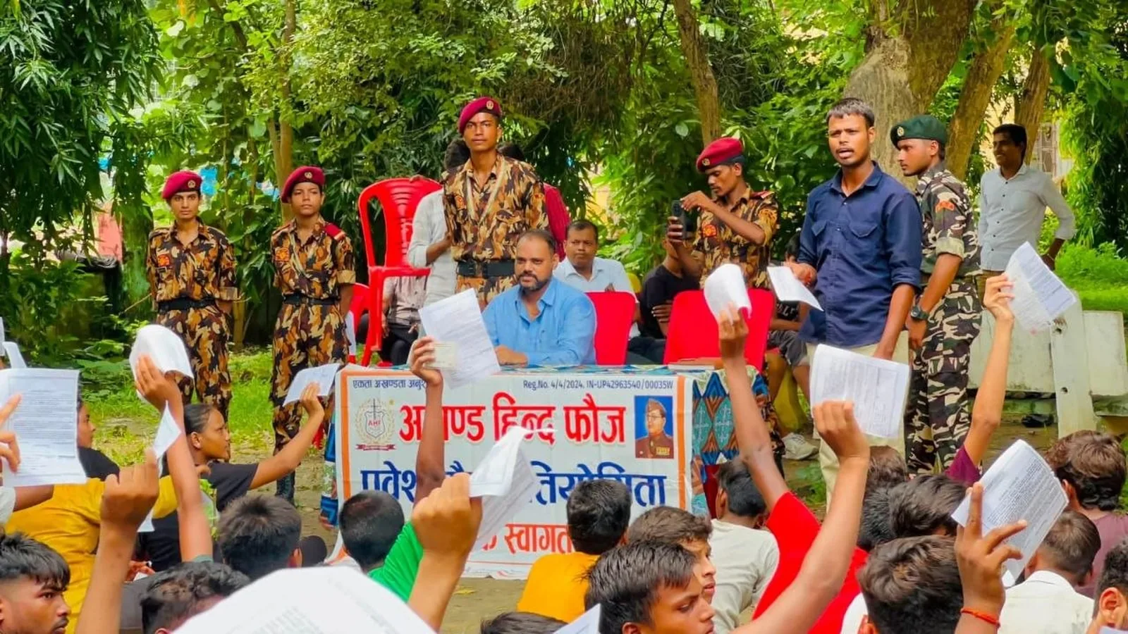 Organisers and uniformed Akhand Hind Fauj members seated behind the event banner as young participants hold printed forms in Pratapgarh