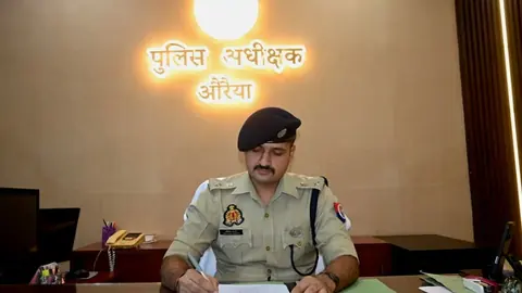 पुलिस अधीक्षक औरैया लिखे रोशन साइन बोर्ड के नीचे मेज पर काम कर रहे वर्दीधारी पुलिस अधिकारी