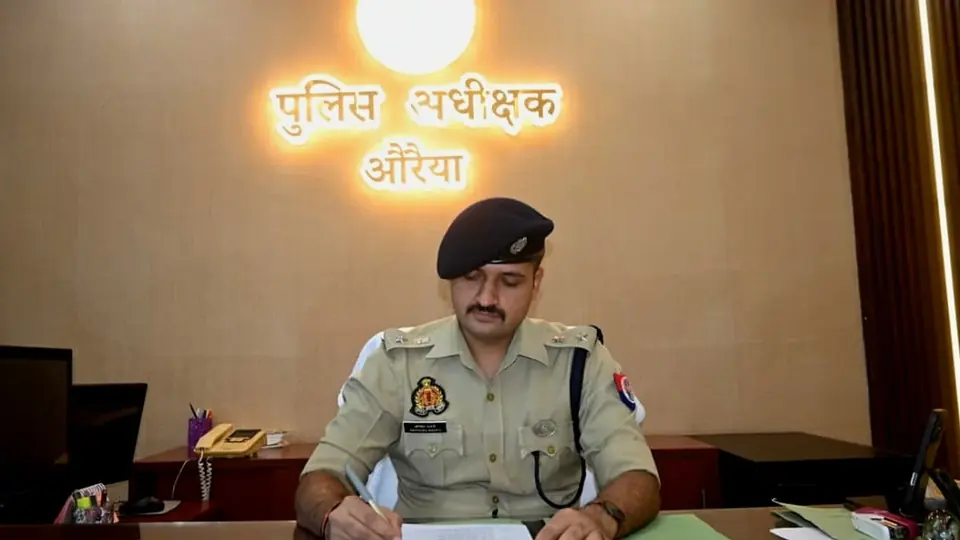 A uniformed police officer works at a desk under an illuminated wall sign reading Superintendent of Police, Auraiya