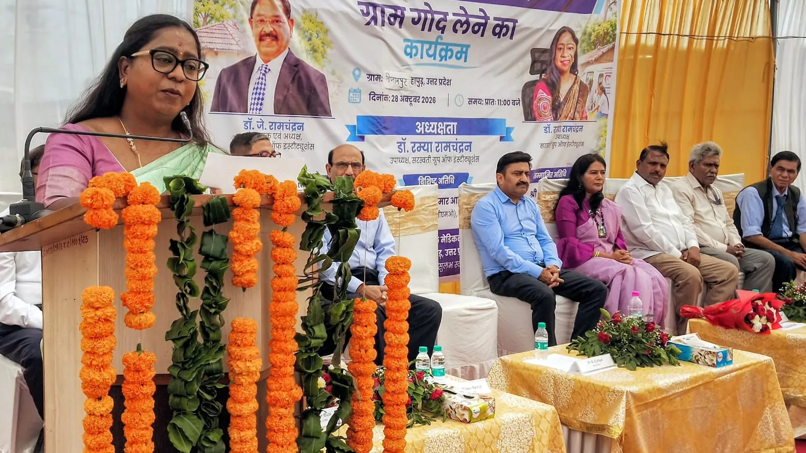 हापुड़ में सरस्वती इंस्टीट्यूट ऑफ मेडिकल साइंसेज के ग्राम गोद लेने के कार्यक्रम को संबोधित करती वक्ता, मंच पर अतिथि मौजूद