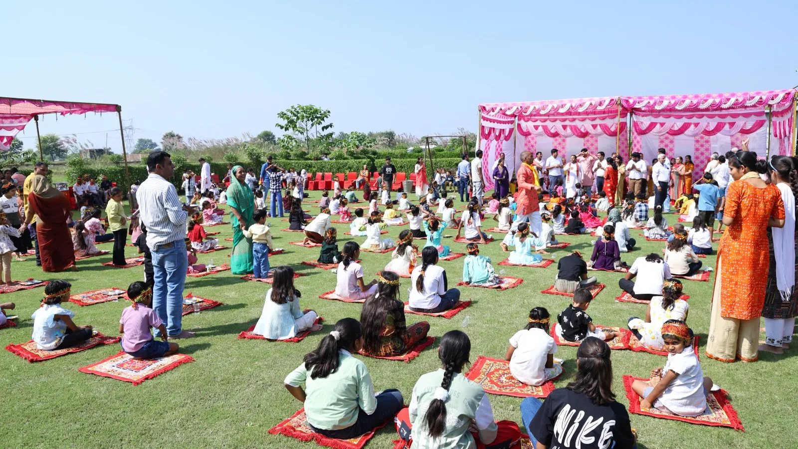 Children seated on red mats on a green lawn with adults nearby at the Sanskar Upvan gathering
