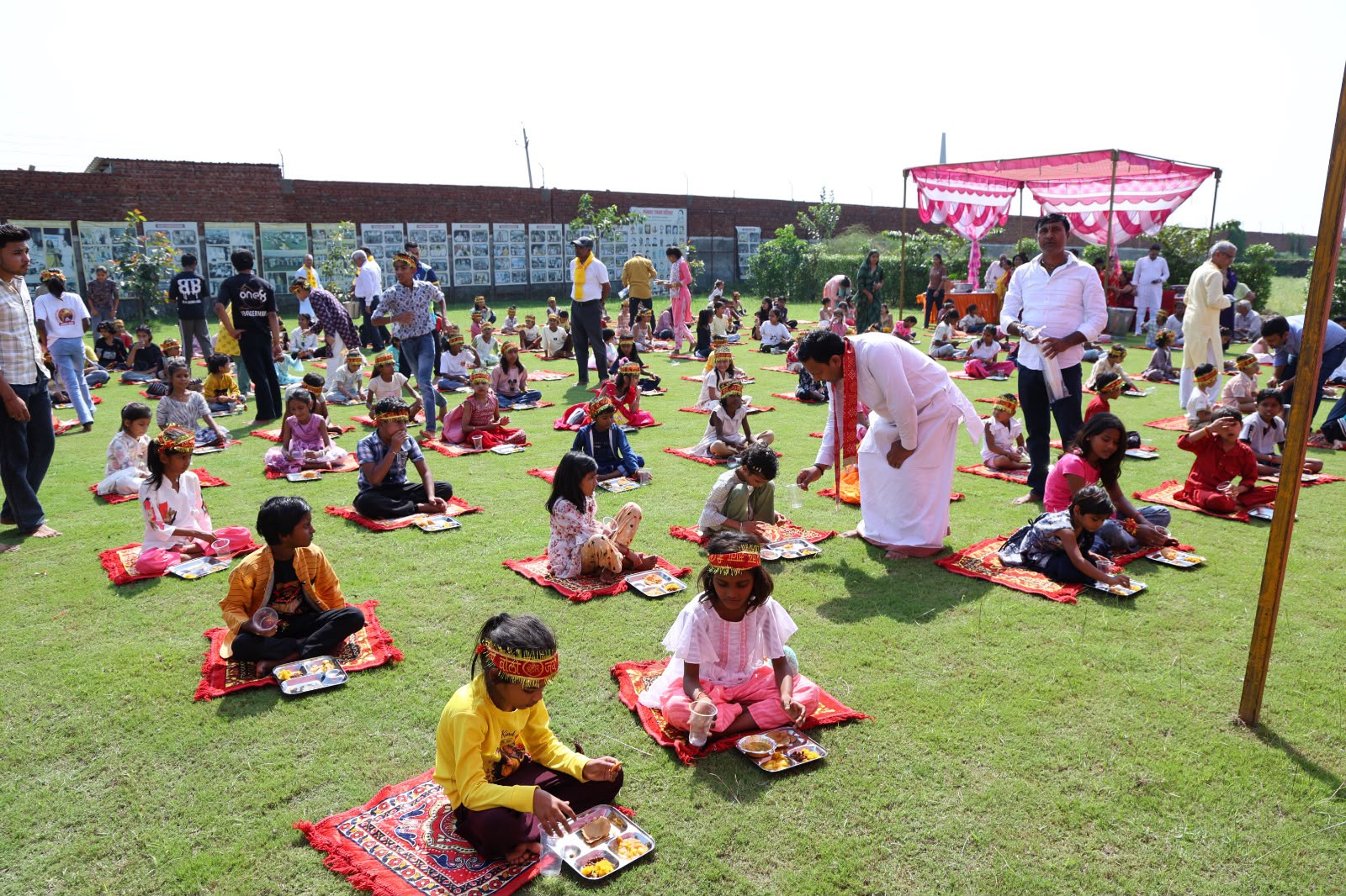 लॉन पर चटाइयों पर बैठकर थालियों में भोजन करते बच्चे और पंक्तियों के बीच वयस्क