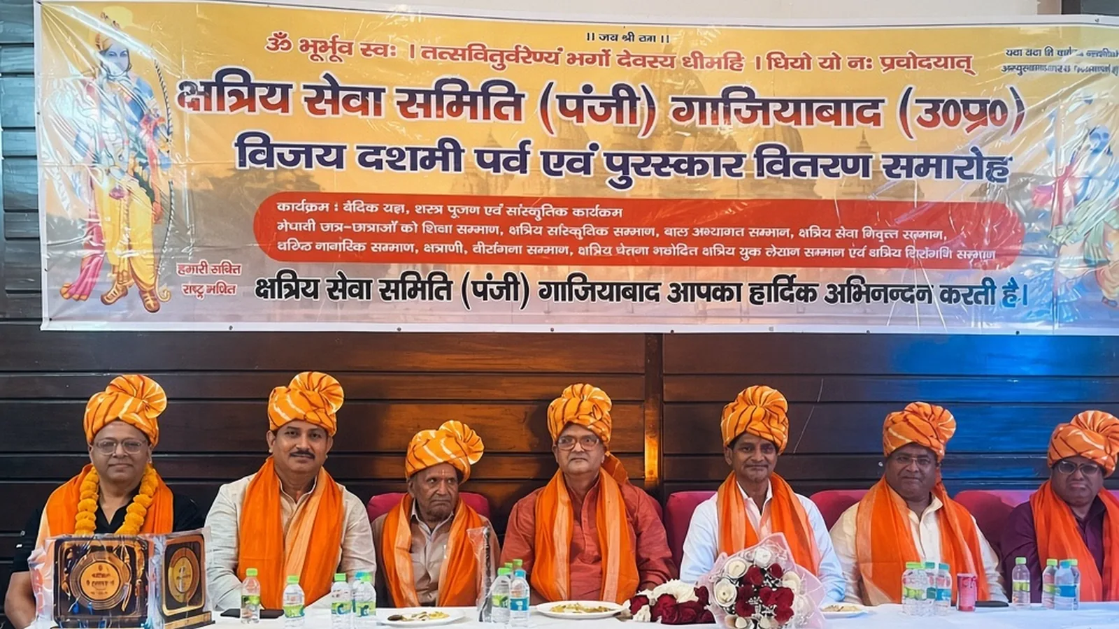 क्षत्रिय सेवा समिति के समारोह में मंच पर नारंगी पगड़ी पहने बैठे अतिथि और सामने रखे पुरस्कार