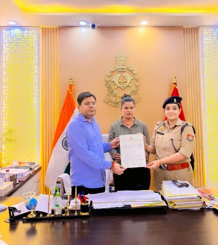 कार्यालय में मेज के पीछे एक दस्तावेज थामे तीन लोग, जिनमें एक महिला खाकी पुलिस वर्दी में हैं