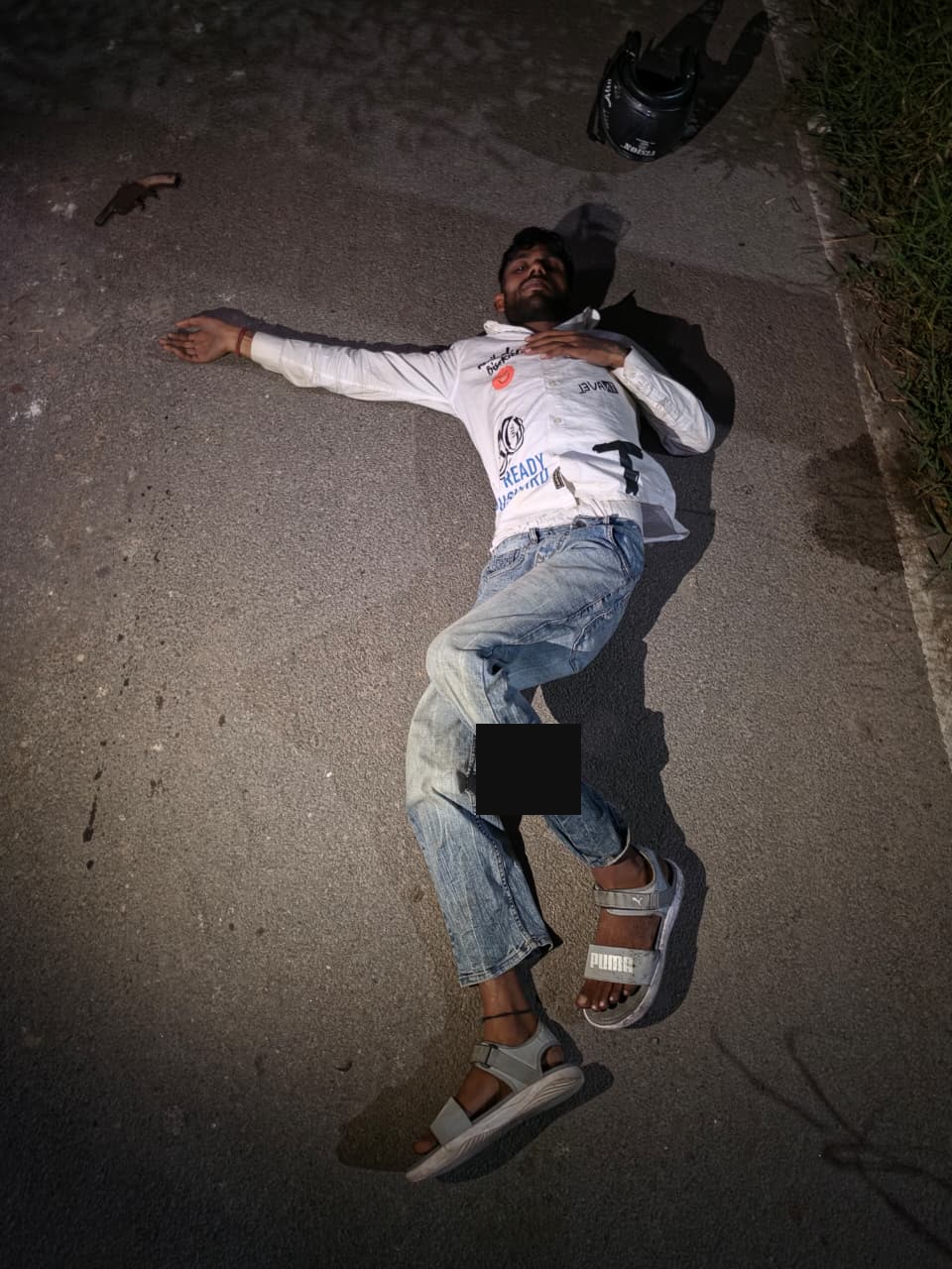 A person on a road at night beside a helmet; the lower-leg area is masked.